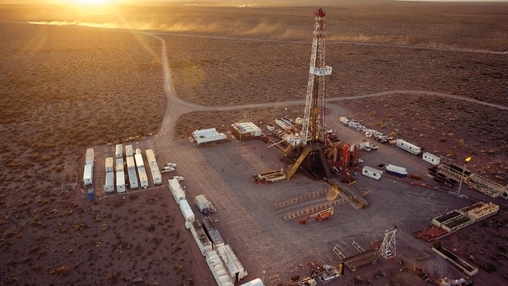 Overhead view of a drilling operation on arid terrain at sunset with equipment yards and a central derrick