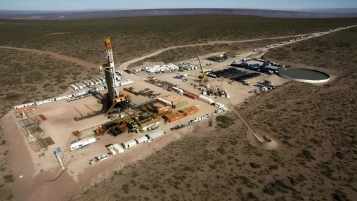 Aerial view of a shale drilling site in arid scrubland with a tall drill rig and a circular water storage pond