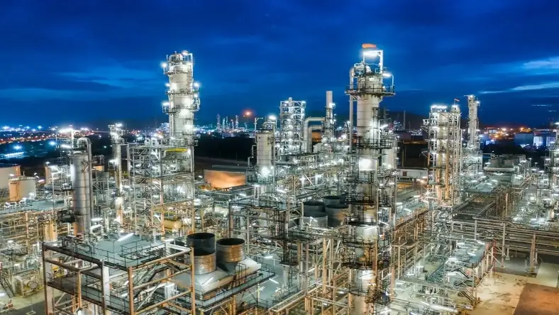 Aerial view of an illuminated oil refinery at night with distillation towers and pipelines against a blue sky