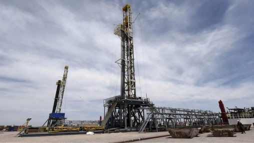 A tall drilling rig with yellow accents stands beside a smaller rig and rows of rusted mud tanks below